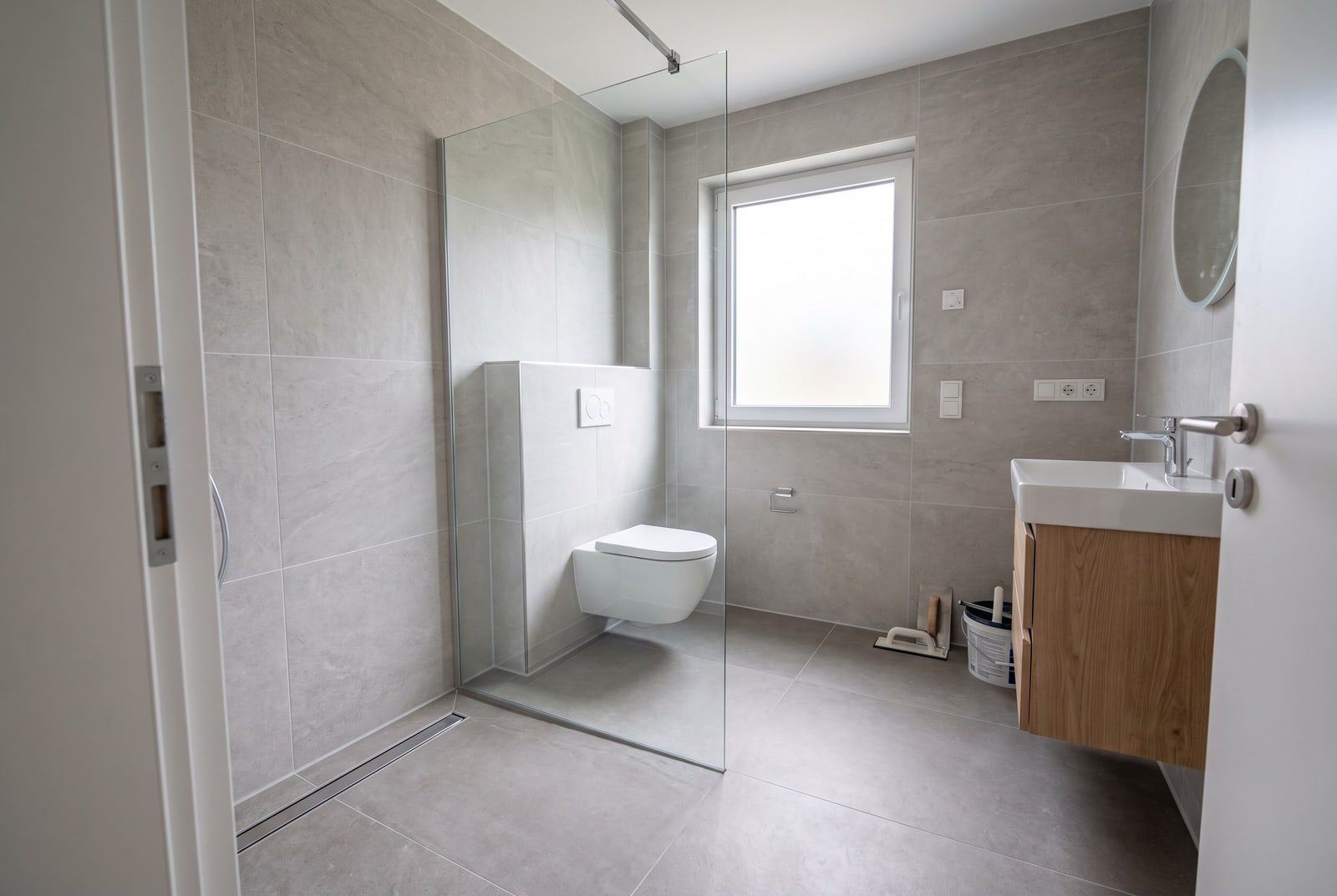 Frisch gefliestes Bad mit großformatigen hellgrauen Fliesen, bodengleicher Dusche mit Glaswand, Hänge-WC und Waschtisch mit Holzunterschrank
