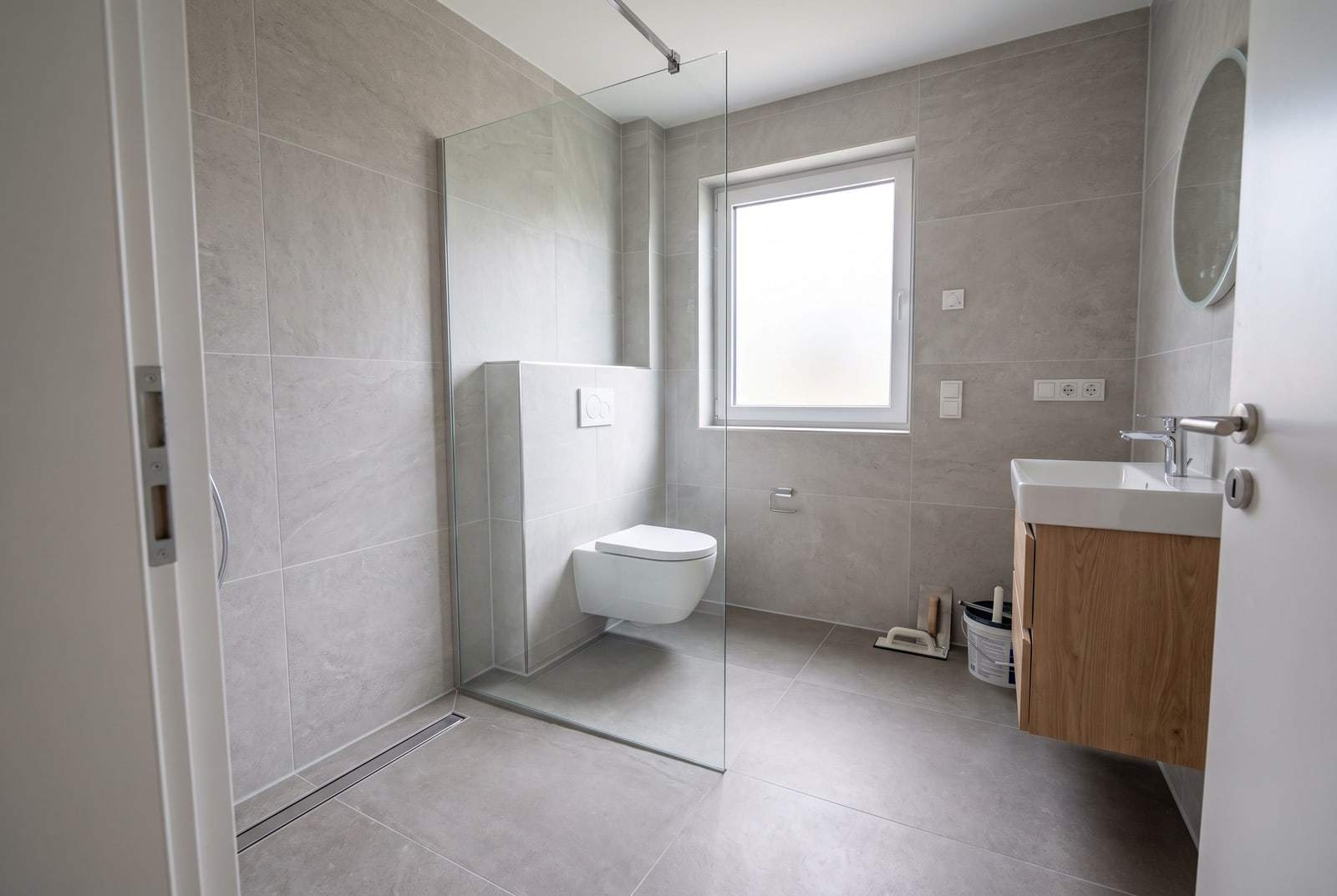 Frisch gefliestes Bad mit großformatigen hellgrauen Fliesen, bodengleicher Dusche mit Glaswand, Hänge-WC und Waschtisch mit Holzunterschrank