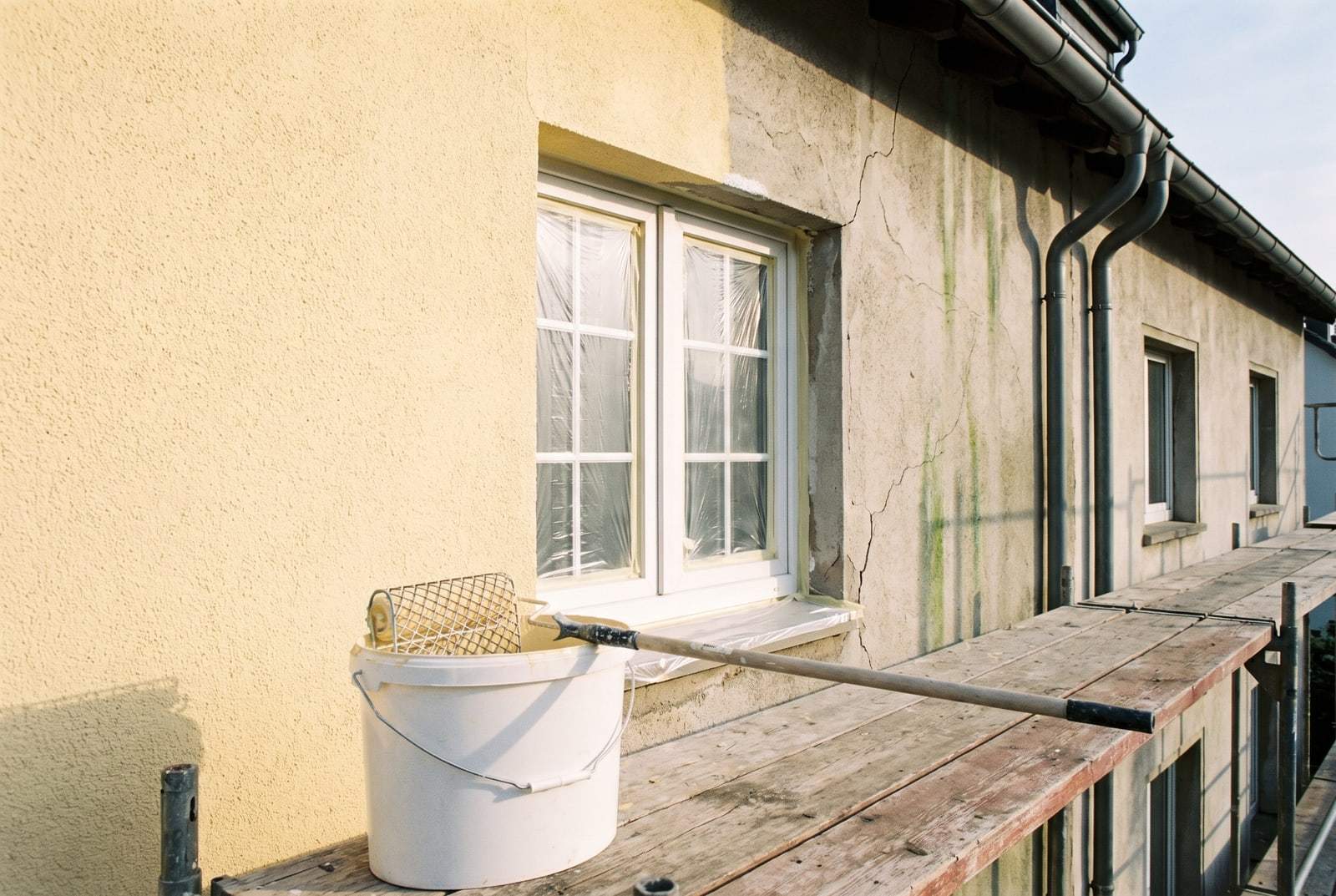 Hausfassade beim Neuanstrich: links frisch hellgelb gestrichen, rechts alter Anstrich mit Algenspuren, Fenster abgeklebt, Farbeimer auf dem Gerüst