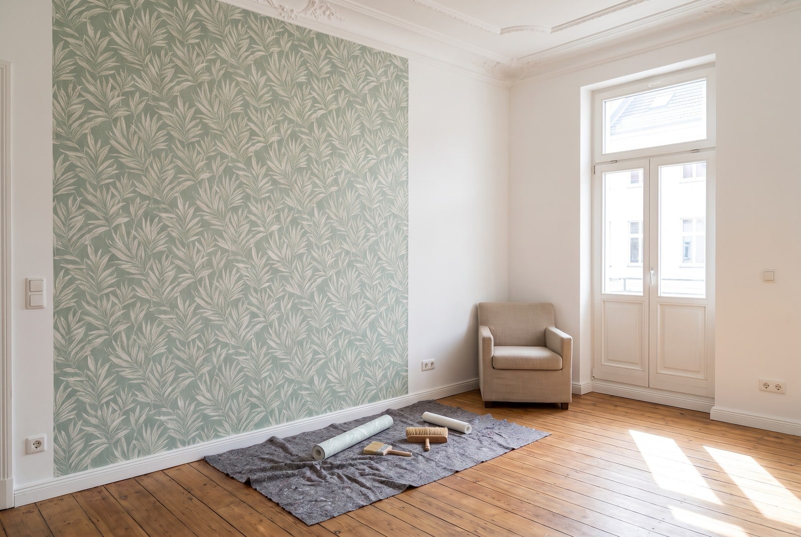 Altbauzimmer mit frisch tapezierter Wand, Vliestapete mit grünem Blattmuster, Tapetenrolle und Bürste auf Malervlies